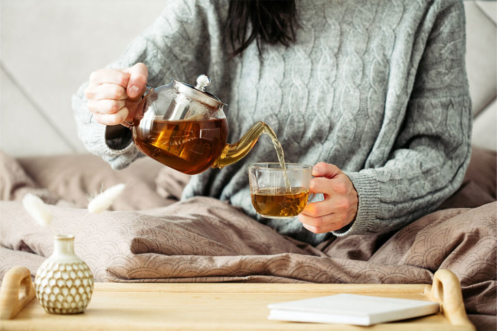 Woman dressed in knitted sweater pours hot tea from glass teapot into mug