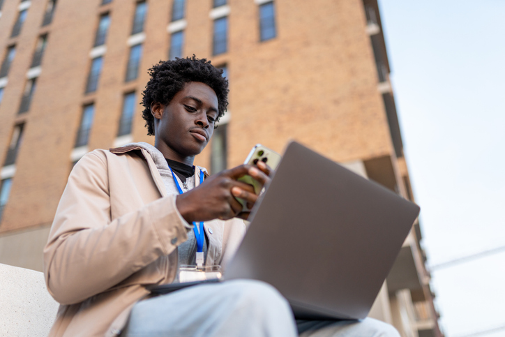 Young adult man sitting outdoors, balancing his laptop and looking at his smartphone, staying connected
