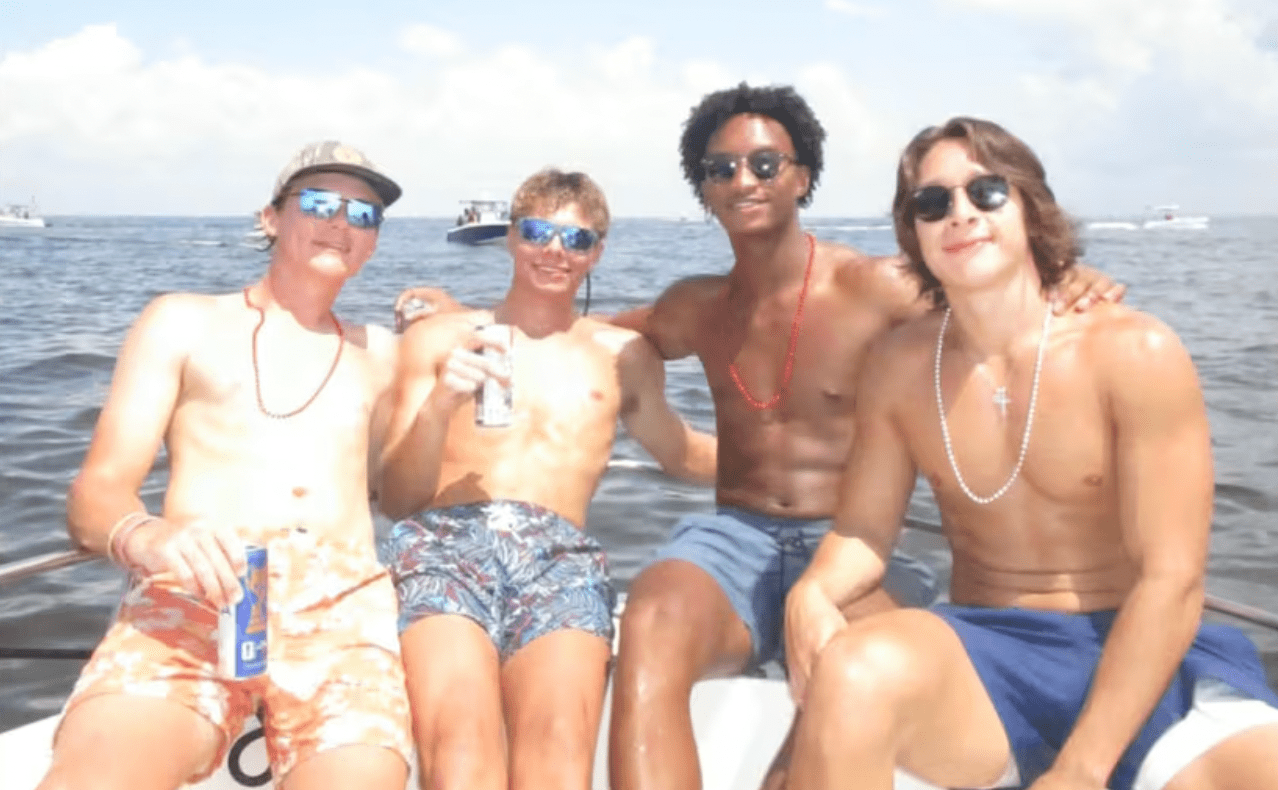 Four young people sitting on a boat in the ocean, wearing sunglasses and swimwear, smiling and enjoying the sunny day.