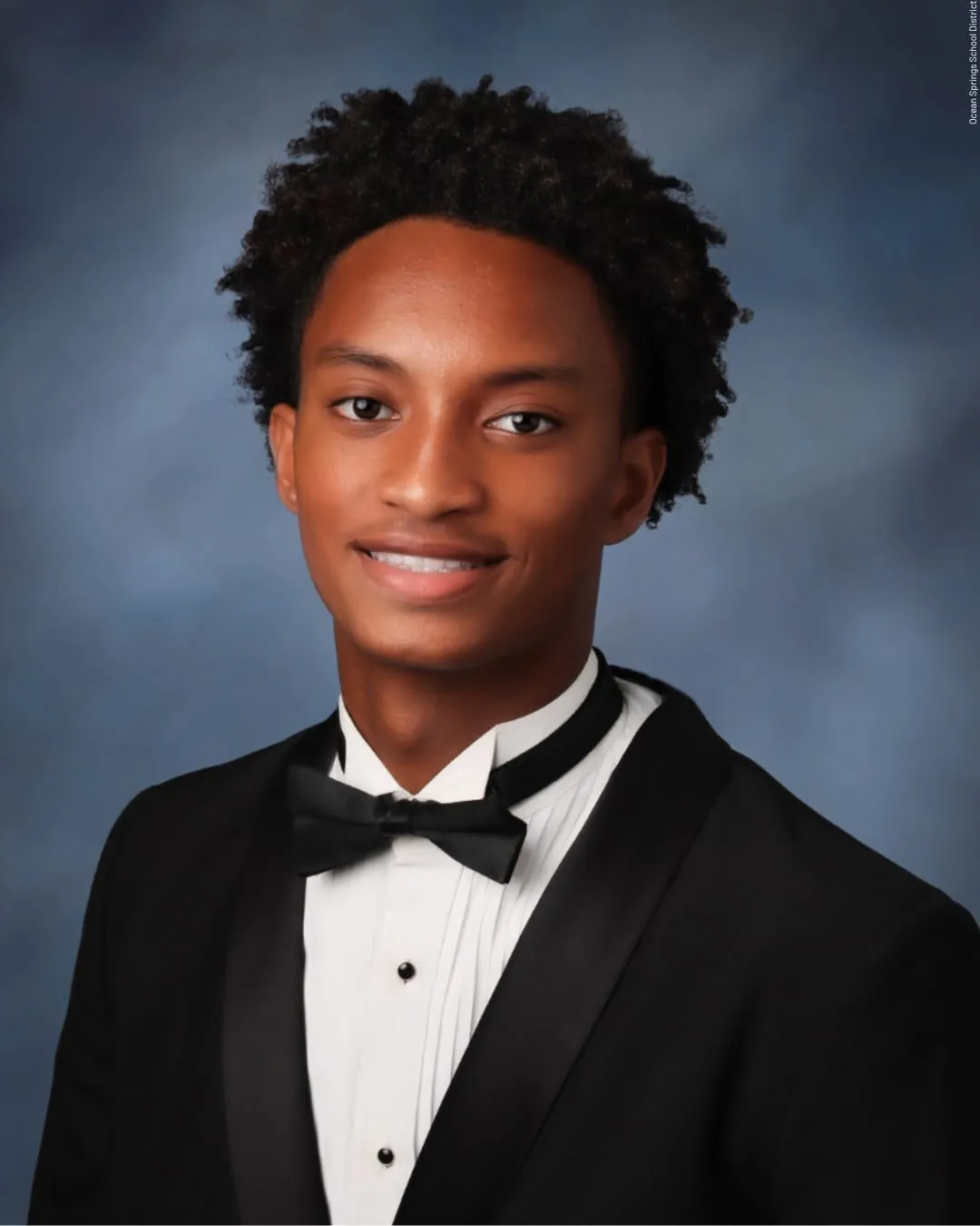 A young Black man wearing a tuxedo with a bow tie, smiling at the camera against a dark blue background.