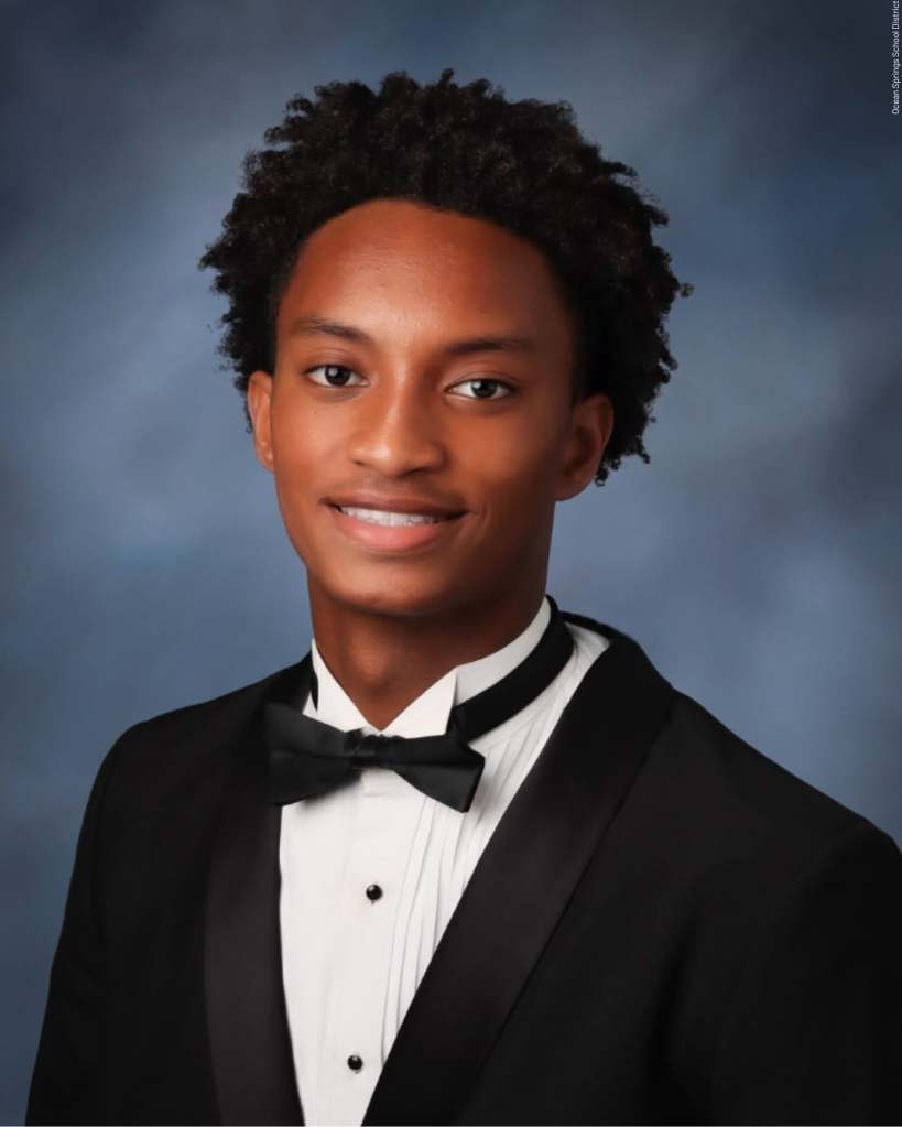 A young Black man wearing a tuxedo with a bow tie, smiling at the camera against a dark blue background.