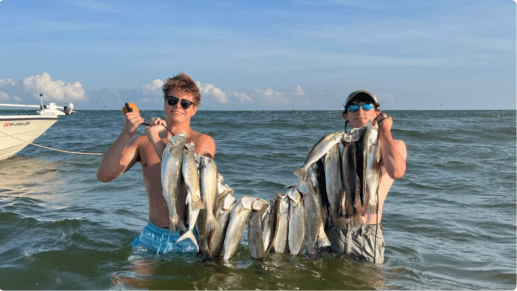 Two men holding up a large catch of fish in the ocean with a boat in the background.