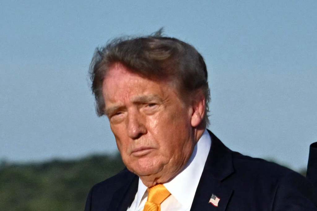 A serious-looking man with dark hair wearing a suit and tie, standing outdoors against a blue sky.