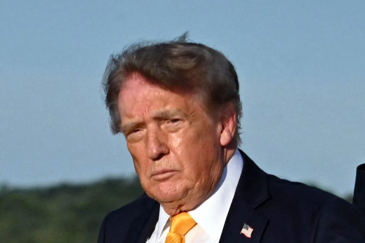 A serious-looking man with dark hair wearing a suit and tie, standing outdoors against a blue sky.