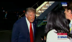 A man in a suit and red tie, identified as Donald Trump, speaking to a reporter in front of a C-SPAN camera and Dallas news ticker.