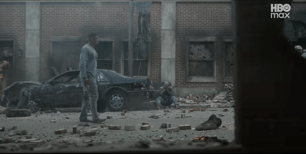 A desolate urban scene with a damaged car and scattered debris on the ground, with a person standing amidst the destruction.