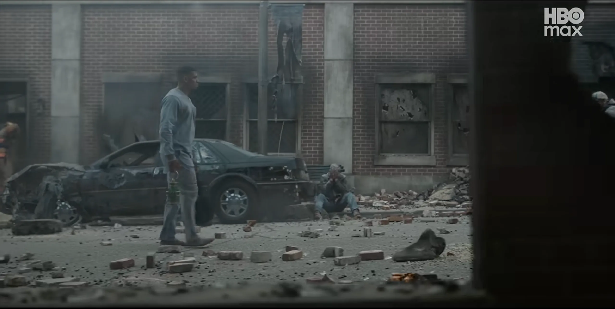 A desolate urban scene with a damaged car and scattered debris on the ground, with a person standing amidst the destruction.