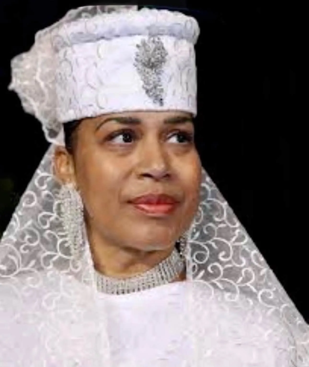 A woman wearing a white headscarf and dress, looking directly at the camera with a serious expression.