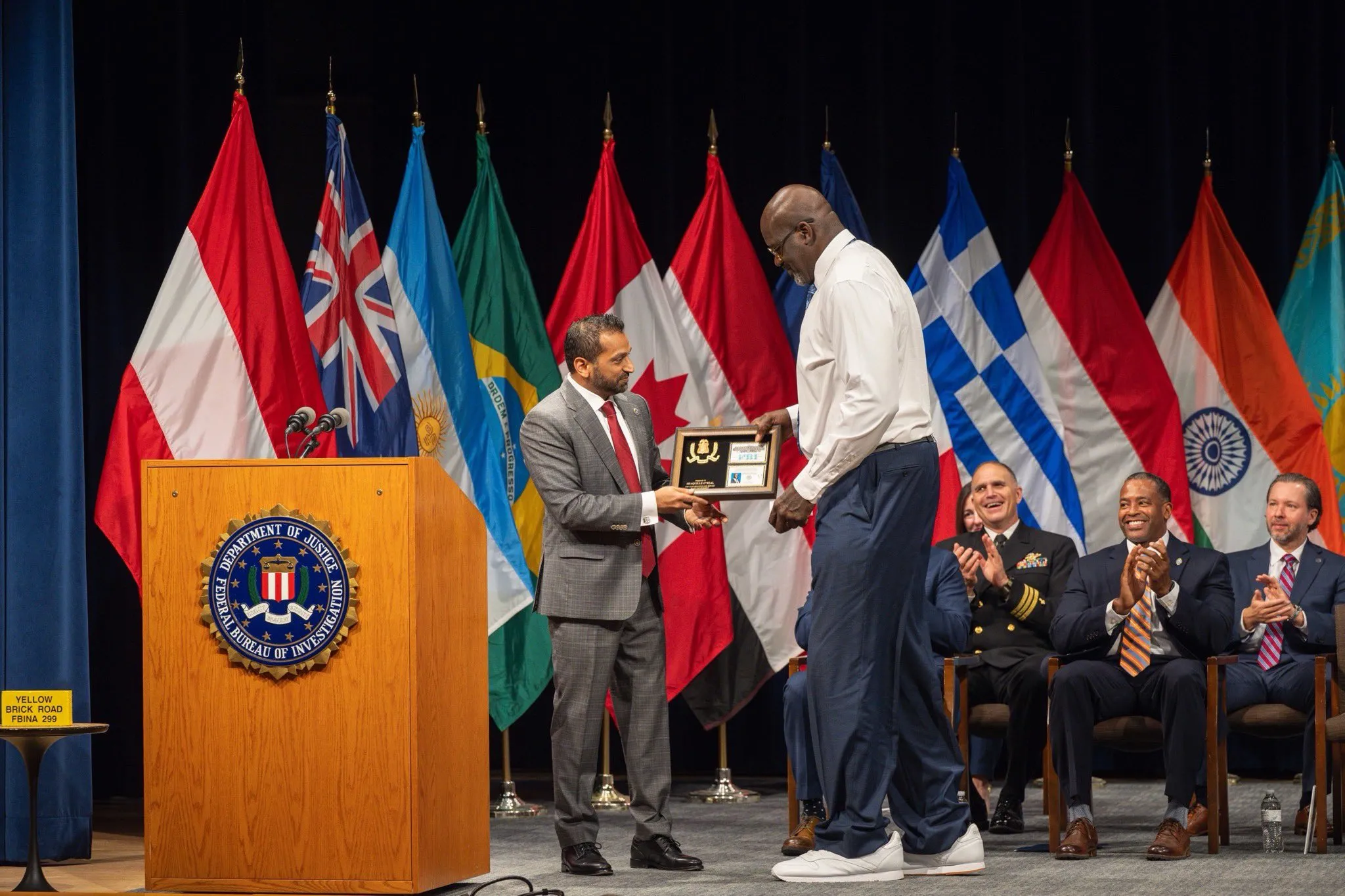 A man in a suit presenting an award to a taller man in a white suit, surrounded by flags and officials applauding.