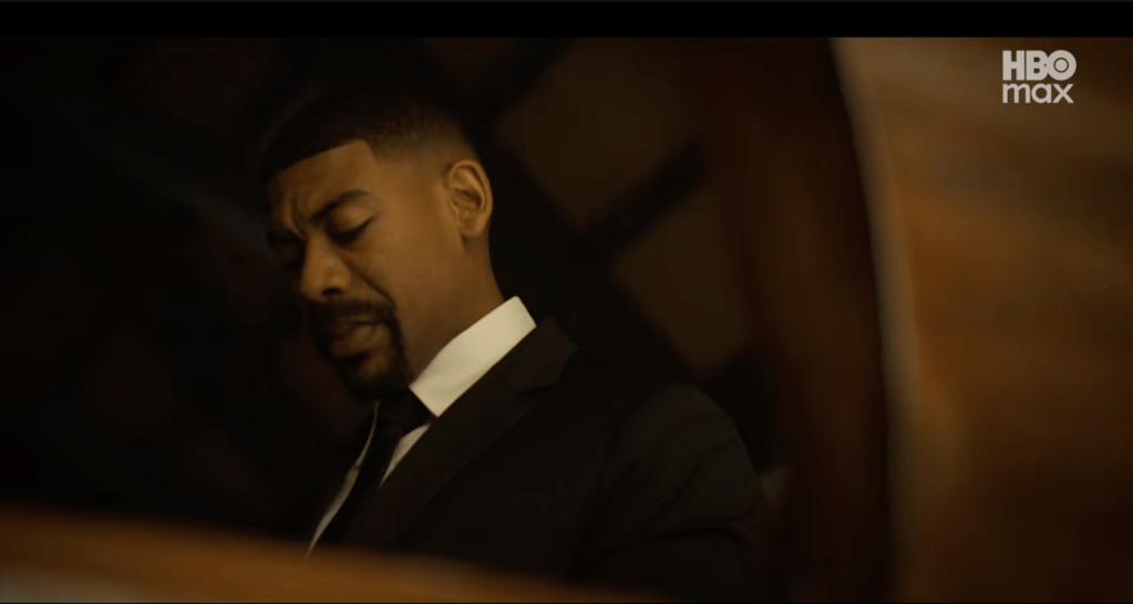 A man in a dark suit with his eyes closed, standing in a dimly lit room.
