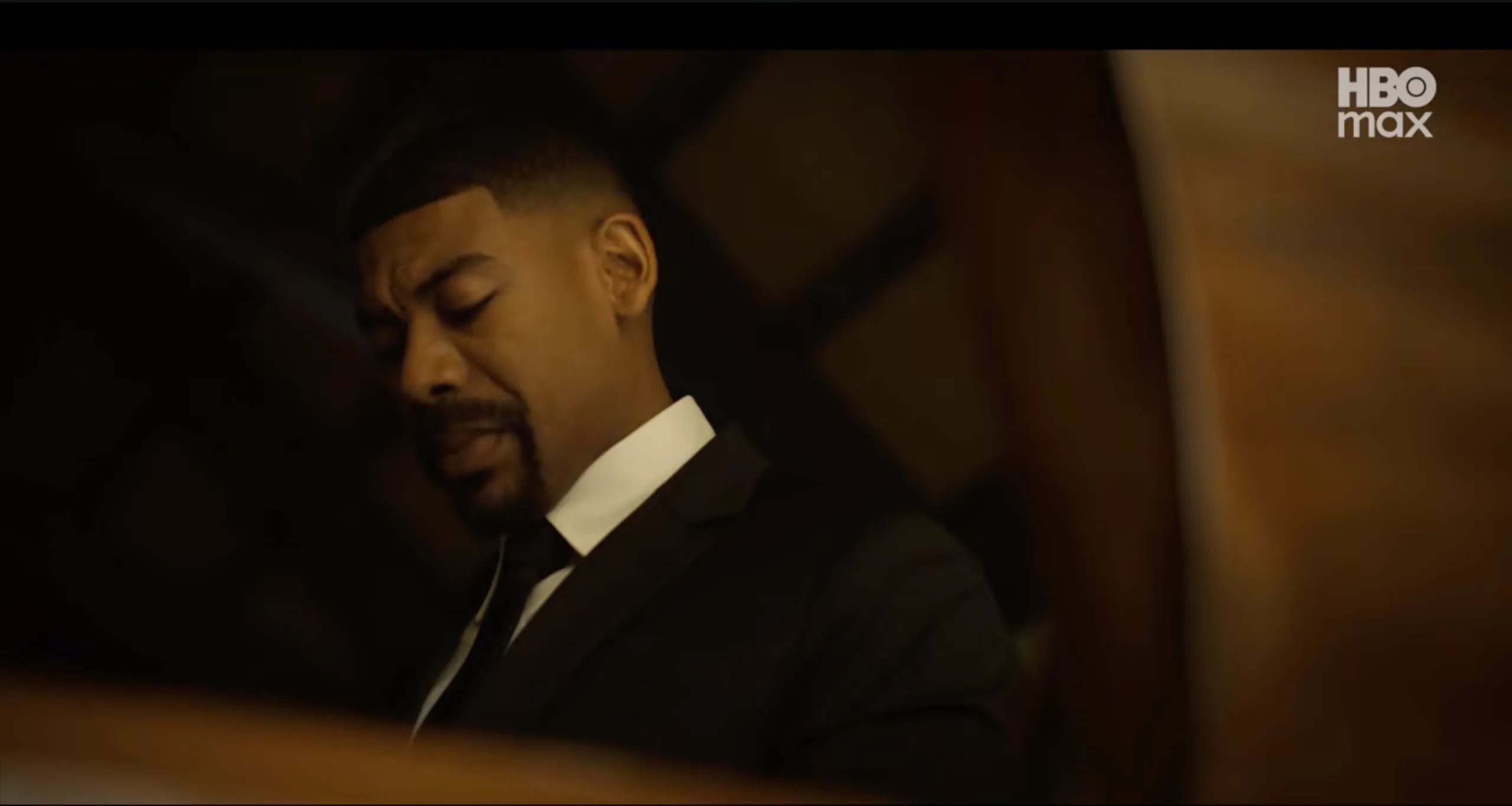 A man in a dark suit with his eyes closed, standing in a dimly lit room.