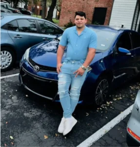 A man in a blue shirt and jeans standing next to a blue Toyota Corolla with damage to the front end.