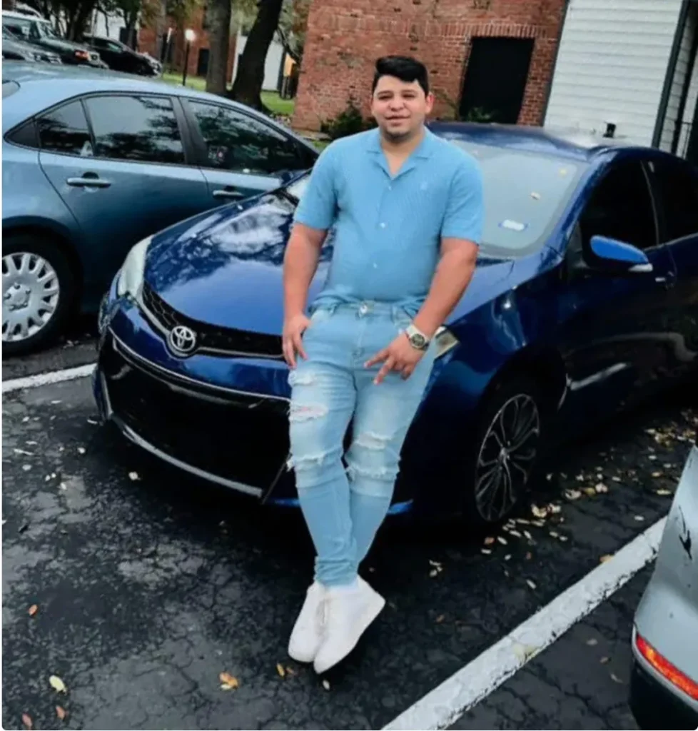 A man in a blue shirt and jeans standing next to a blue Toyota Corolla with damage to the front end.