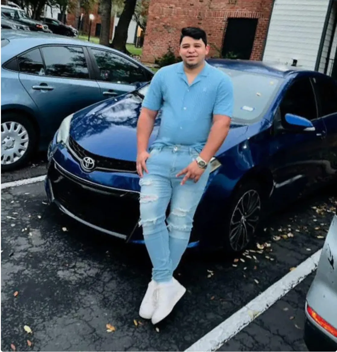 A man in a blue shirt and jeans standing next to a blue Toyota Corolla with damage to the front end.