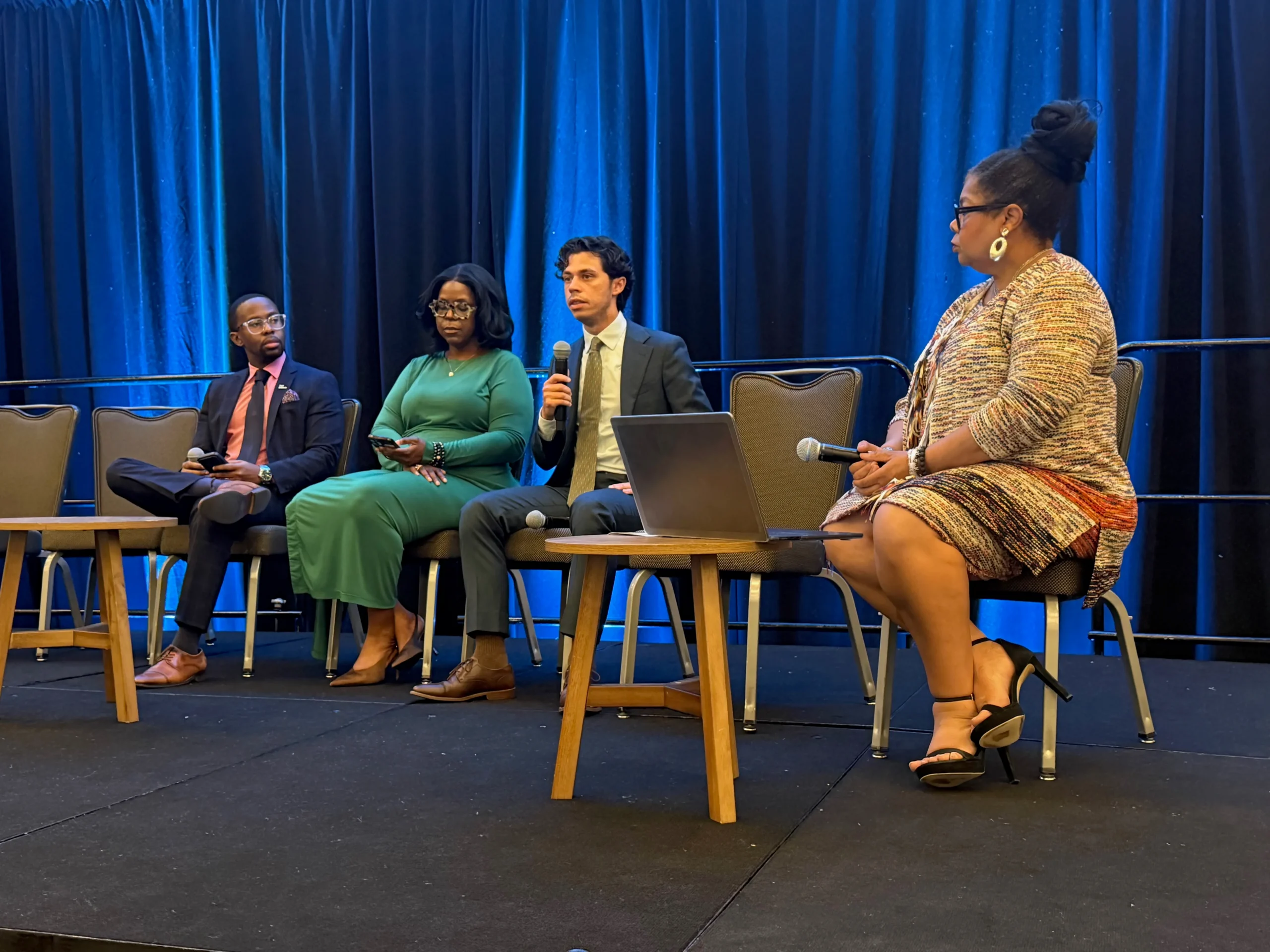 Four panelists seated on stage, discussing a topic. The woman in the center is speaking into a microphone, while the other three panelists listen. The backdrop is a blue curtain.