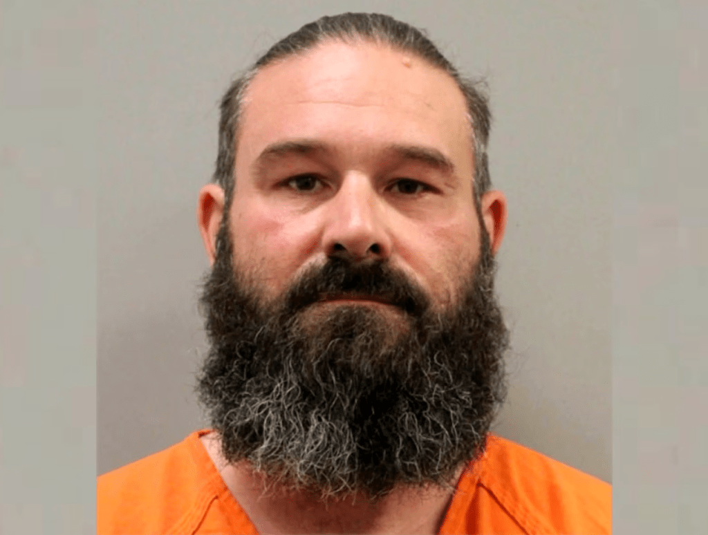 Mugshot of a man with a long beard wearing an orange prison uniform.