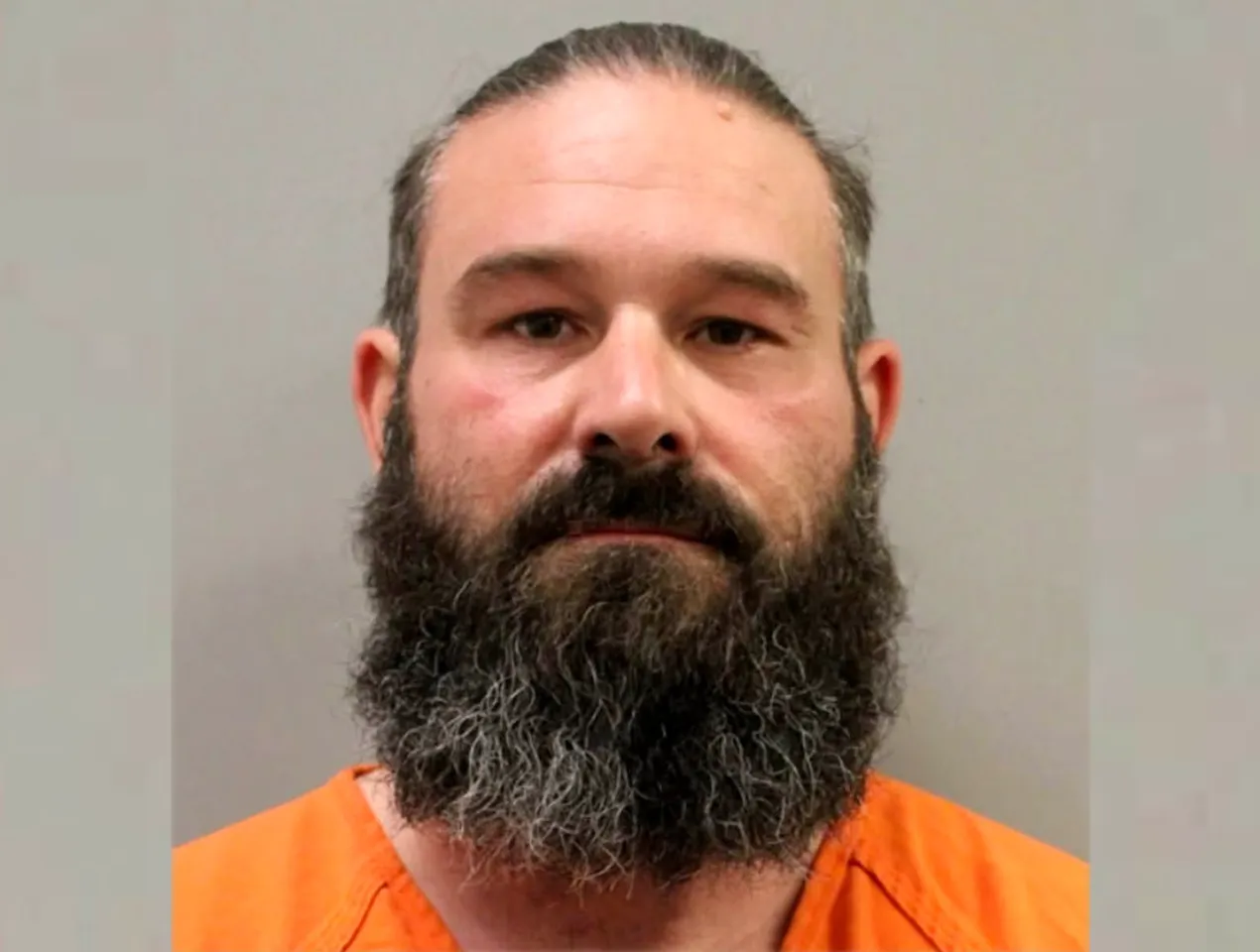Mugshot of a man with a long beard wearing an orange prison uniform.