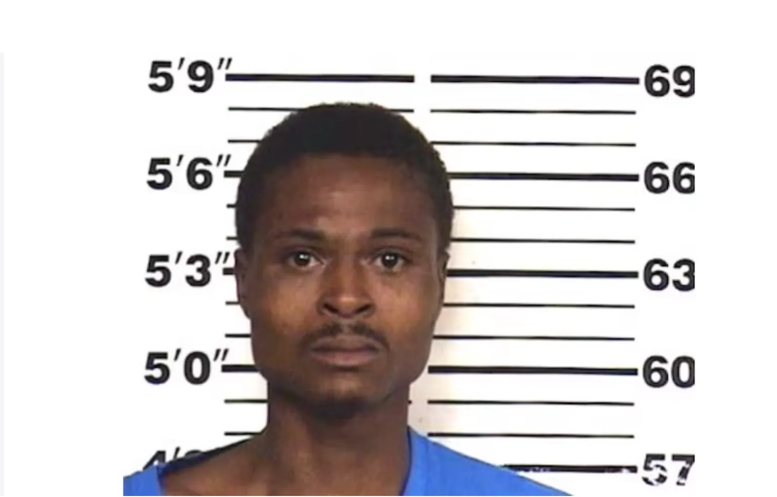 Mugshot of a man with a serious expression, wearing a blue shirt, against a height measurement backdrop.