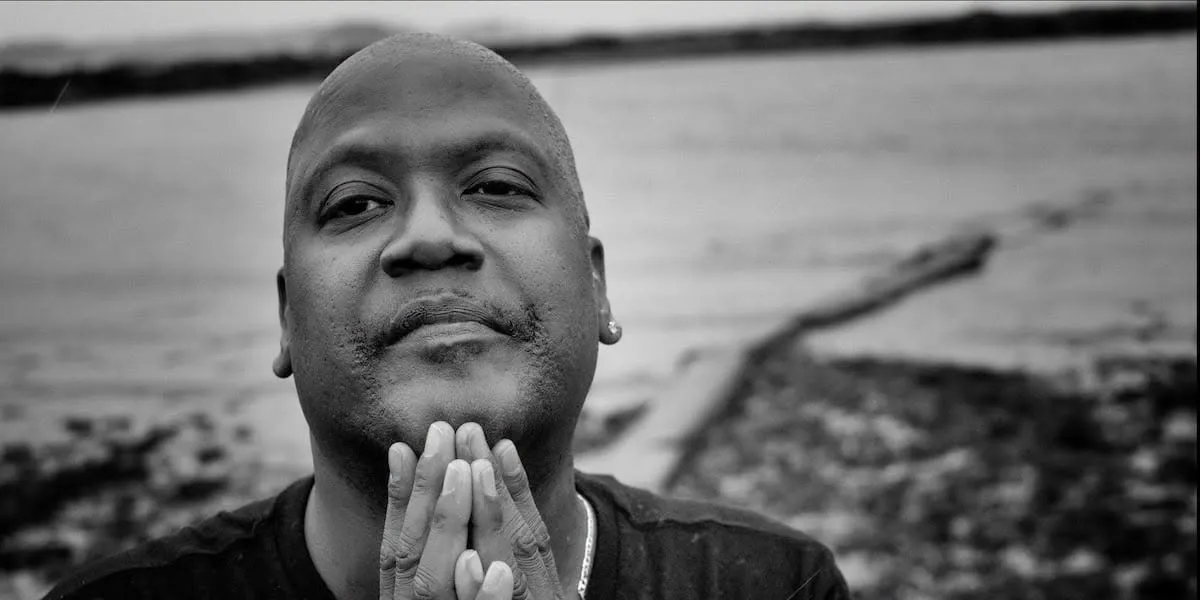 Black and white portrait of Curtis Flowers, a man with a shaved head, hand raised to his chin in a contemplative pose, with a body of water and rocky shoreline visible in the background.