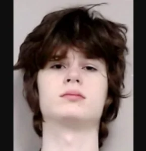 Mugshot of a young man with shoulder-length dark brown hair and a neutral expression against a light gray background.