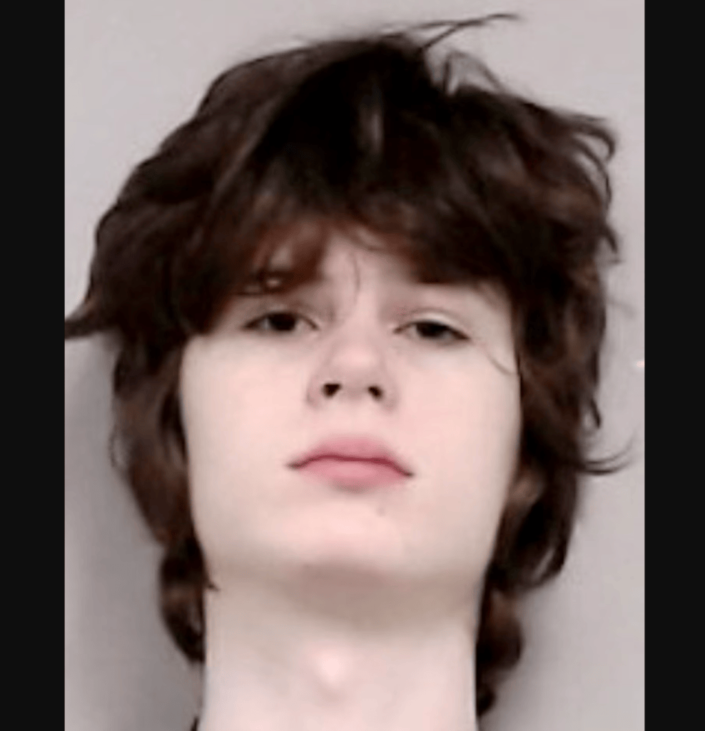Mugshot of a young man with shoulder-length dark brown hair and a neutral expression against a light gray background.