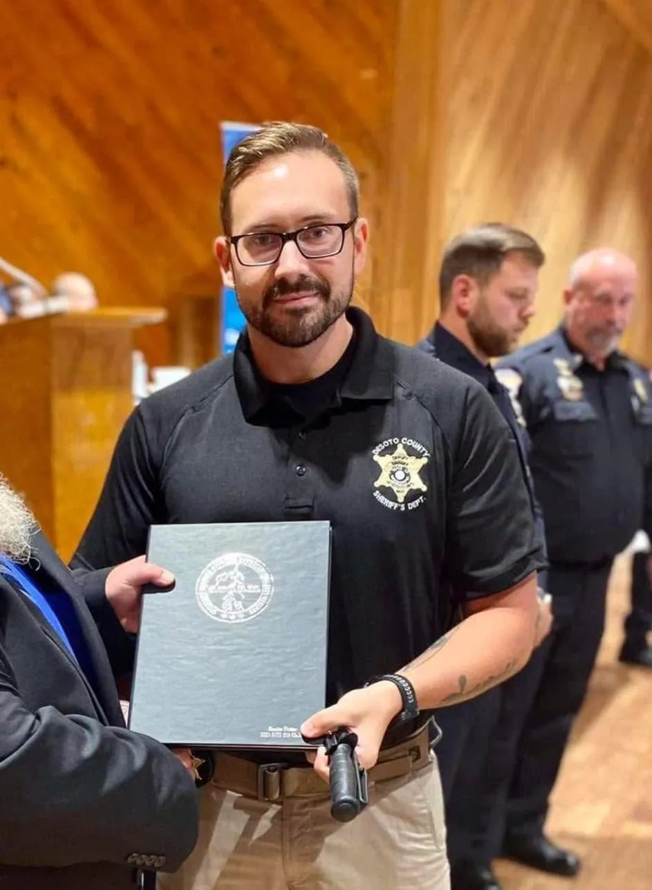 A man wearing glasses and a black Senatobia Police Department polo shirt holds a light blue certificate or award folder during an indoor ceremony, with other uniformed officers visible in the background.