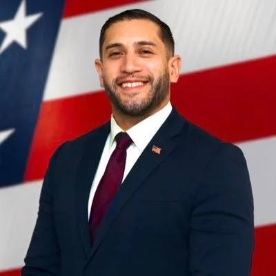 Professional headshot of a man in a dark suit and maroon tie smiling at the camera against an American flag background.